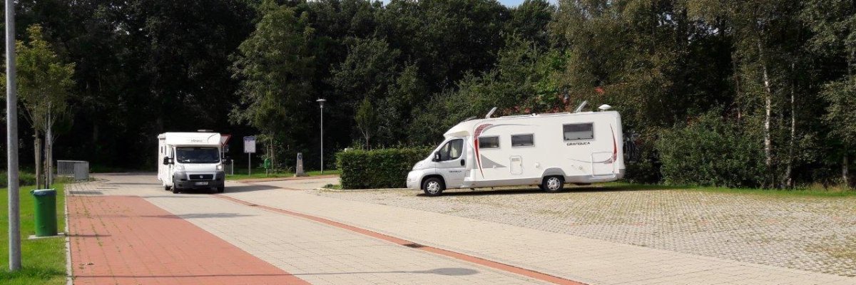 Auf dem Bild sieht man den Hager Wohnmobilstellplatz, auf dem ein Wohnmobil steht und einer kommt angefahren. Umgeben ist der Platz mit vielen großen Bäumen.