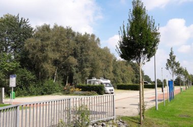 Auf dem Bild sieht man den Wohnmobilstellplatz von der Einfahrt aus. Im rechten Vordergrund ist ein Metallzaun und im Hintergrund sieht man die Stellplätze mit Bäumen ganz hinten. Auf dem Stellplatz steht ein Wohnmobil.