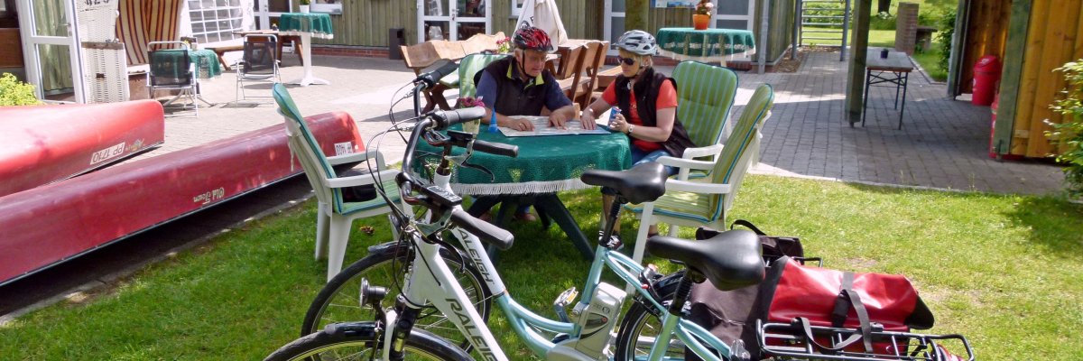 Zwei Fahrräder mit Gepäck stehen an der Paddel und Pedal Station auf dem Rasen. im Hintergrund sieht man einen kleinen Baum und auf dem Boden liegende Kanus. Ganz hinten steht ein kleinen Häuschen, in dem sich die Station befindet.