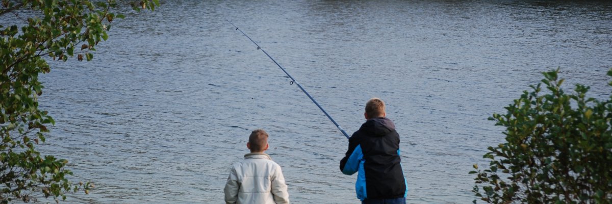 Zwei Jungs mit heller und dunkler Jacke. Mit dem Rücken zur Kamera in Richtung See. Der eine Junge hat eine Angel ausgeworfen.
