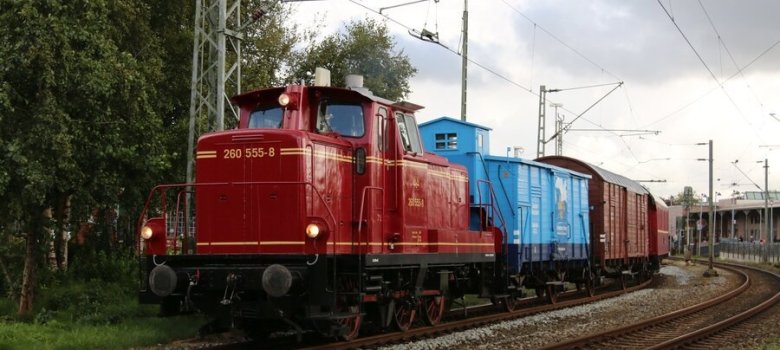 Auf dem Bild sieht man eine Museumseisenbahn in Rot. der zweite Waggon ist hellblau. Rechts sieht man weitere Schienen und einen Bahnhof. links neben dem Zug sind Bäume. 