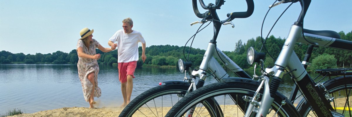 Im Vordergrund befinden sich zwei Fahrräder, die an einem kleinen Strand am See stehen. Die Sonne scheint und im Hintergrund ist ein lachendes Pärchen, das auf die Fahrräder zurennt. 