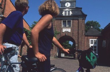 Vor dem Tor des Lütetsburger Schlosses stehen, mit dem Rücken zum Bild, zwei Frauen mit dem Fahrrad und schauen sich das Schloss an. 