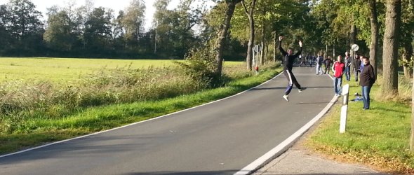 Auf dem Bild erkennt man eine lange graue Landstraße. Links und rechts davon sind Rasen und Grünflächen und es stehen Bäume an den Seiten. Auf der Straße ist ein Mann, der eine Boßelkugel wirft und dafür in die Luft springt. Am Rand stehen andere Personen, die ihm zugucken. 