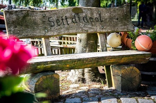 Auf dem Bild erkennt man eine alte Holzbank mit der Innschrift "Sett di daal". Links erkennt man verschwommen eine rosa Blume und rechts von der Bank ist Dekoration aus einem Holzfass und Bojen. 