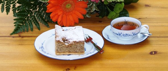 Auf dem Bild sieht man einen Kuchenteller und eine Teetasse. Auf dem Kuchenteller ist ein Stück Kuchen mit Puderzucker und einer kleinen Gabel. In der Tasse ist bereits Tee und das Geschirr passt zusammen. Beides in weiß mit dunkel Blauer Verzierung. Dahinter liegt ein Blumenstrauß mit einer Orangenen Blüte. 