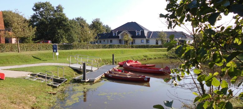 Auf dem Bild sieht man einen Anleger im Hager Tief, an dem 4 Kanus / Kajaks liegen und ein Tretboot. Alle Boote sind rot. links sieht man viel Rasenfläche und im Hintergrund ein großes Haus mit Weisem Backstein. 
