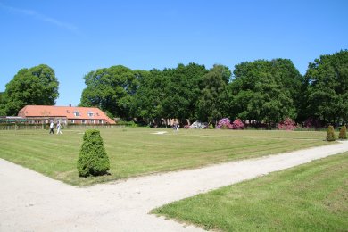 Auf dem Bild ist ein Teil des Lütetsburger Schlossparks zu sehen. Im Hintergrund befindet sich das Schlosscafe, mit einem Hellen Dach. Davor befindet sich eine große Rasenfläche mit einen Kiesweg, der sich im Vordergrund des Bildes befindet. Rechts neben dem Cafe sieht man viele dichte, dunkelgrüne Bäume.