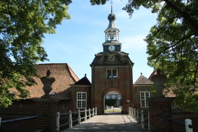 Auf dem Bild erkennt man den Eingang zum Lütetsburger Schloss. Es ist eine Brücke im Vordergrund, die zu einem Turm mit Eingang führt. Oben drauf ist eine Kuppel mit einer Uhr und links und rechts geht das Gebäude weiter.
