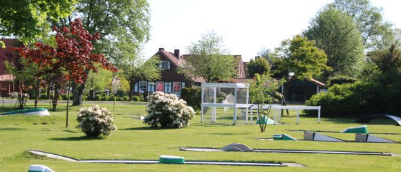 Auf dem Bild sieht man eine Minigolfanlage, die auf einer Rasenfläche ist. Im Hintergrund sieht man Bäume und Büsche und ein Wohnhaus.