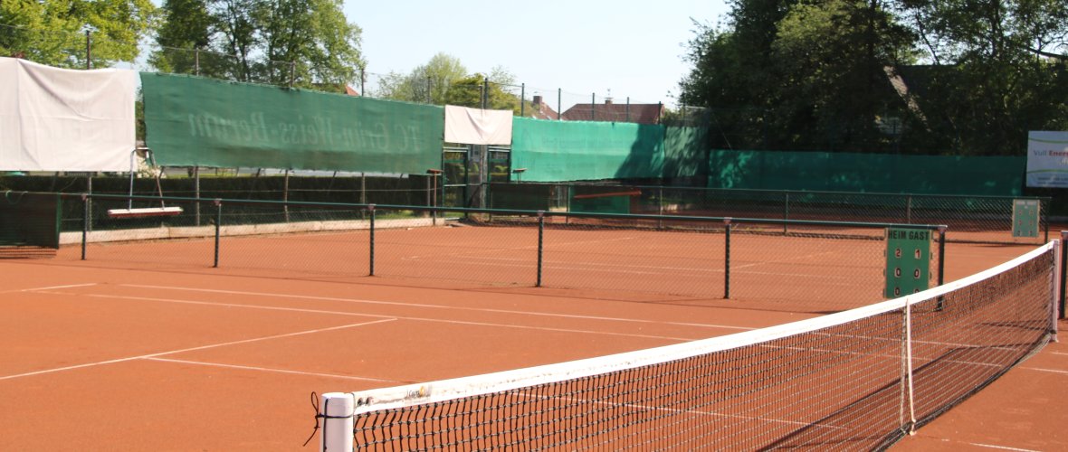 Auf dem Bild sieht man einen Tennisplatz, der von der seitlichen Mitte aus fotografiert wurde. Dort ist ein Netz zusehen und der Boden ist in einem Orange-braun. 