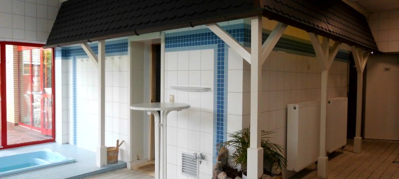Auf dem Bild ist eine Sauna von außen zu sehen. diese ist mit weißen Fliesen bedeckt und davor steht ein weißer Stehtisch. Links erkennt man ein kleines Bad vor einem Fenster. 