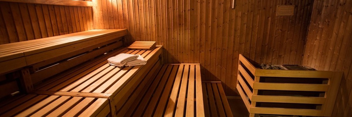 Auf dem Bild ist das innere einer Sauna zu sehen. Es sind drei Holzbänke und rechts ein Ofen. Auf der mittleren Bank liegt ein Handtuch. 