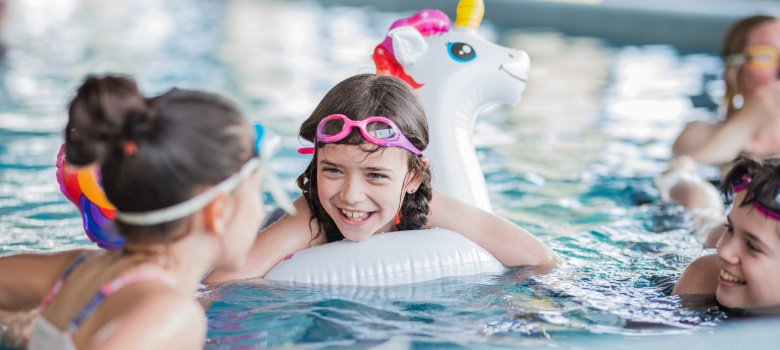 Happy girl floating in swimming pool on inflatable unicorn and talking to her friends