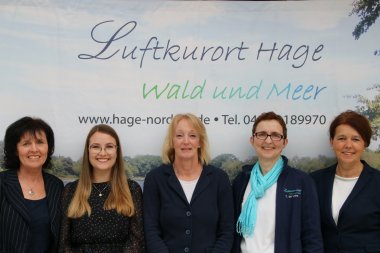 Auf dem Bild ist das Team der Kurverwaltung Hage zu sehen. Es sind 5 Frauen vor einer Leinwand mit dem Luftkurort Hage Logo.